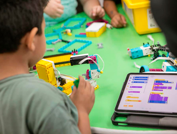 Niño arma un robot de lego en una mesa con más niños, junto a una tablet que muestra información. 