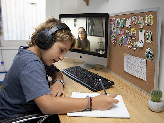 Niño escribe sobre un cuaderno usando audífonnos y junto a un computador donde se ve una mujer.