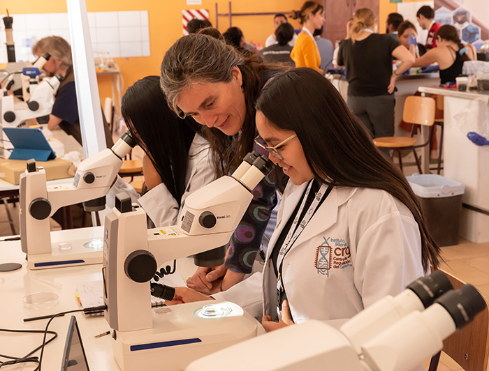 Una estudiante observa por microscopio junto a una profesora, en un laboratorio.