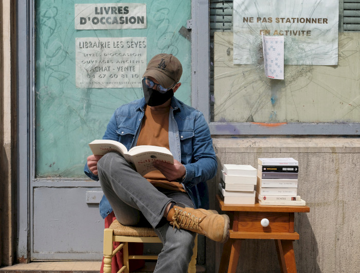 Hombre con mascarilla sentado en una vereda leyendo libros.