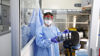 Fotografía de una mujer en un laboratorio, con todos los equipos de protección personal, que sostiene una serie de tuvos de ensayo.