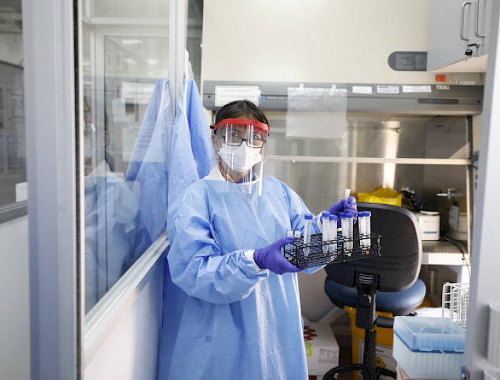 Fotografía de una mujer en un laboratorio, con todos los equipos de protección personal, que sostiene una serie de tuvos de ensayo.