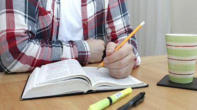 imagen de persona con lápiz sobre libro en escritorio donde hay más útiles y una taza