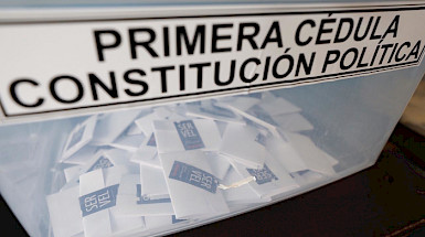 Fotografía que muestra una urna de las elecciones del Plebiscito 2020 en Chile. Por fuera de la caja de plástico se lee: Primera cédula, constitución política. Dentro de la caja transparente se ven varios votos emitidos y cerrados con un adhesivo del SERVEL.