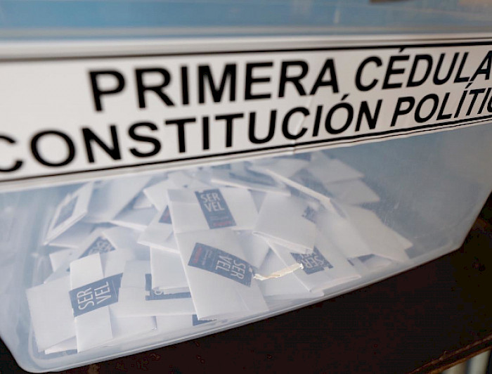 Fotografía que muestra una urna de las elecciones del Plebiscito 2020 en Chile. Por fuera de la caja de plástico se lee: Primera cédula, constitución política. Dentro de la caja transparente se ven varios votos emitidos y cerrados con un adhesivo del SERVEL.