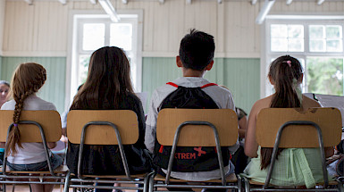 Niños y niñas sentados en sillas de colegios y vistos desde la espalda.