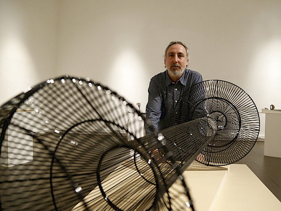 Escultura metálica, tubular, de pintura negra, en primer plano. Detrás, escultor de la obra mirando de frente, en una sala de exposición.