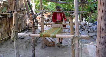 Fotografía de una mujer manipulando telas e hilos en una estructura de madera, en un espacio abierto y rural donde se ven paredes de maderas, piedras y árboles.