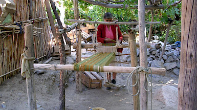 Fotografía de una mujer manipulando telas e hilos en una estructura de madera, en un espacio abierto y rural donde se ven paredes de maderas, piedras y árboles.