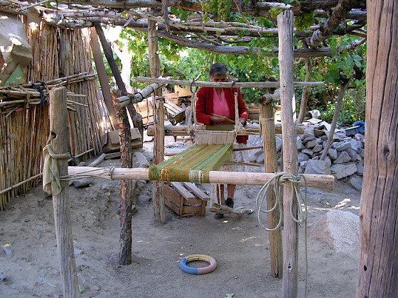 Fotografía de una mujer manipulando telas e hilos en una estructura de madera, en un espacio abierto y rural donde se ven paredes de maderas, piedras y árboles.