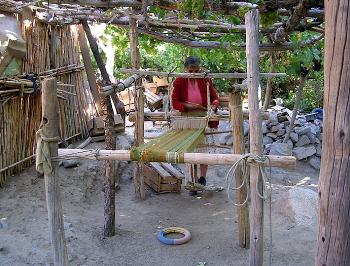 Fotografía de una mujer manipulando telas e hilos en una estructura de madera, en un espacio abierto y rural donde se ven paredes de maderas, piedras y árboles. 