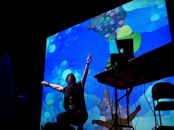 Hombre, sentado, brazos al aire. Al lado hay una mesa con un notebook Apple. Detrás hay una pantalla con gráficas azules.