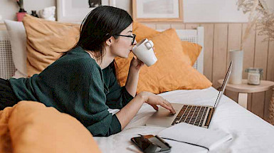 a woman is sitting on her bed with a laptop and a cup of coffee