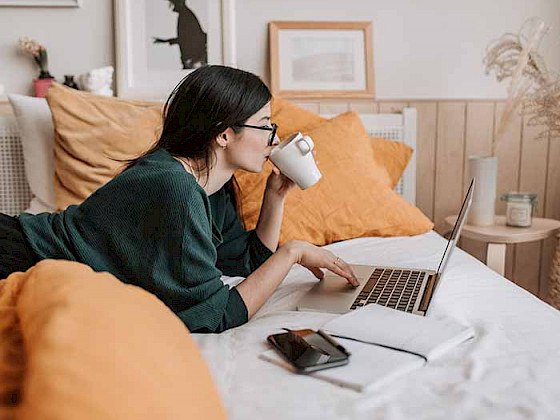 a woman is sitting on her bed with a laptop and a cup of coffee