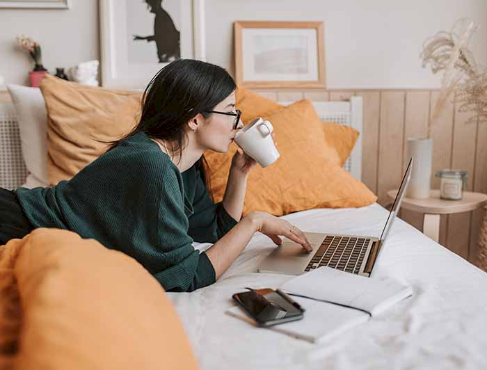a woman is sitting on her bed with a laptop and a cup of coffee