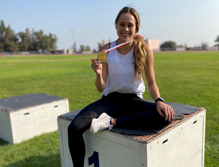 La la estudiante y velocista María Ignacia Montt muestra su medalla de oro sentada en el podio del primer lugar en una cancha de pasto. 