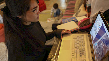 Imagen de joven mamá ante un computador junto a su hijo pequeño que juega en medio del living.