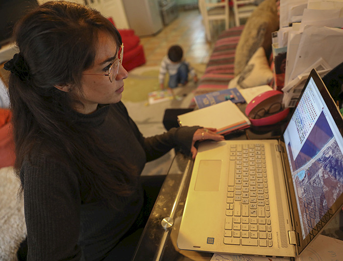 Imagen de joven mamá ante un computador junto a su hijo pequeño que juega en medio del living. 