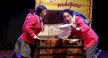 Rosario y Gaspar, dos escolares, sentados frente a un baúl de madera, descubriendo un objeto antiguo. Detrás de ellos, un libro grande dice El Volantín Mágico.