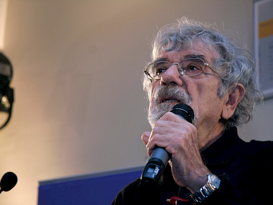 hombre con pelo canoso, barba blanca y anteojos, hablando con un microfono en la mano