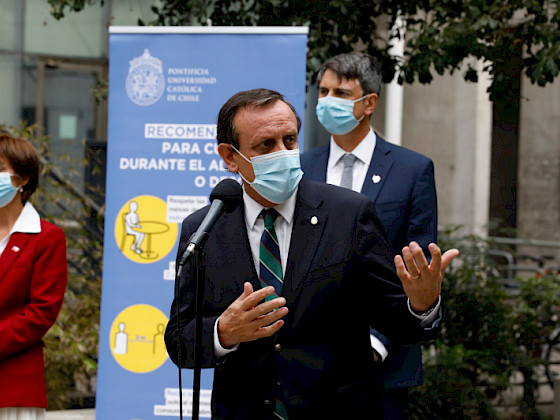 hombre con mascarilla hablando frente a microfono, de pie, personas detras de él