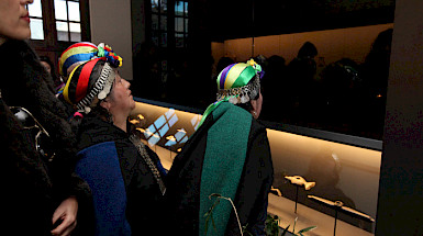 Cuatro mujeres, tres de ellas con la vestimenta tradicional mapuche, admirando la exposición del Aula de Arte Pueblos Originarios, en el Campus Oriente UC.