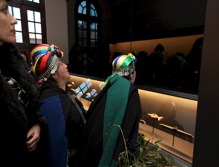 Cuatro mujeres, tres de ellas con la vestimenta tradicional mapuche, admirando la exposición del Aula de Arte Pueblos Originarios, en el Campus Oriente UC. 