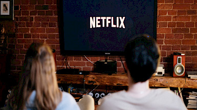 Dos personas de espaldas, sentadas en un sofá, frente a un televisor que en la pantalla tiene el logo de Netflix.