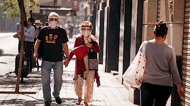 Pareja de adultos mayores con mascarilla y escudo facial caminando por una vereda de Santiago un día soleado.