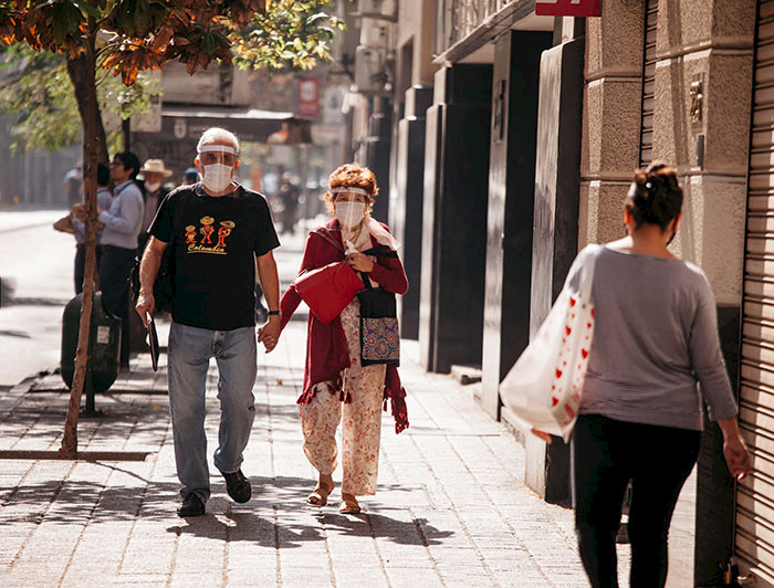 Pareja de adultos mayores con mascarilla y escudo facial caminando por una vereda de Santiago un día soleado.