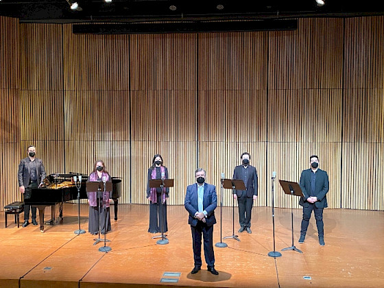 Foto panorámica del escenario del Centro de Extensión Oriente, con cuatro cantantes de pie, con mascarillas y partituras. Además de un pianista y piano de cola en el fondo de la imagen y al centro el director del coro de cámara UC. Todos con ropa de presentación formal.