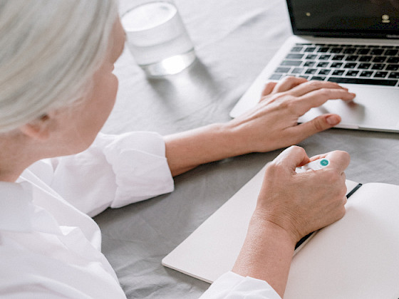 Mujer, pelo blanco, de espaldas, sentada frente a su notebook, junto a una agenda en la que está escribiendo con su mano derecha.