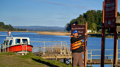 hombre frente a un lago y a una lancha señalando un letrero