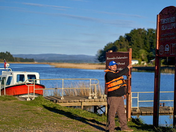 hombre frente a un lago y a una lancha señalando un letrero