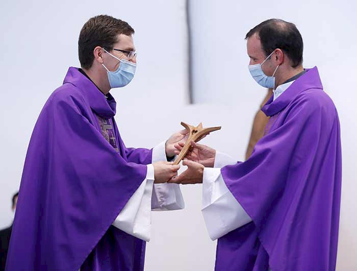 dos sacerdotes, el de la derecha recibe un crucifijo de madera