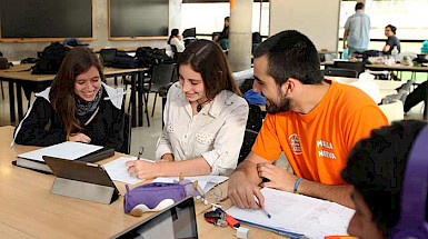 dos estudiantes mujeres y un estudiante hombre sentados alrededor de una mesa con lápices, cuadernos y tablet