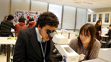 A young man is watching through a microscope, while a woman is supervising him.