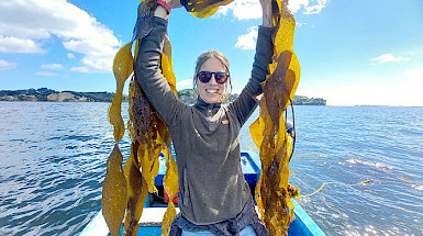 María José de la Fuente is smiling and standing in a boat, while is holding seaweed above his head.