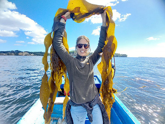 María José de la Fuente is smiling and standing in a boat, while is holding seaweed above his head.