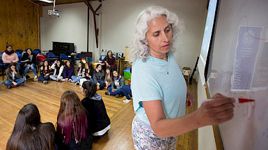 A woman is writing in a whiteboard with a red marker while a group of young women are paying attention to her.