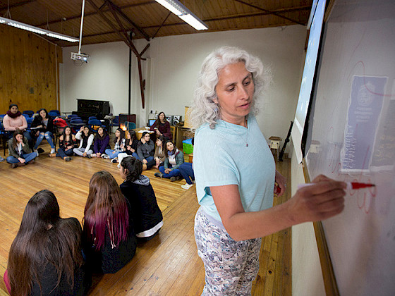 A woman is writing in a whiteboard with a red marker while a group of young women are paying attention to her.