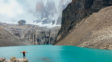 Persona con sus brazos abiertos mira el lago y al fondo las Torres del Paine.