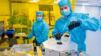 Two scientists wearing masks and blue suits manipulate elements in the laboratory.