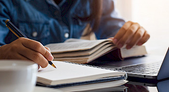 Mujer frente a un computador escribiendo en un cuaderno, junto a un libro y computador.