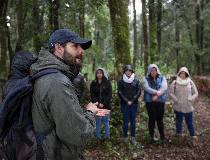 A man is talking in front of six people in a forest.