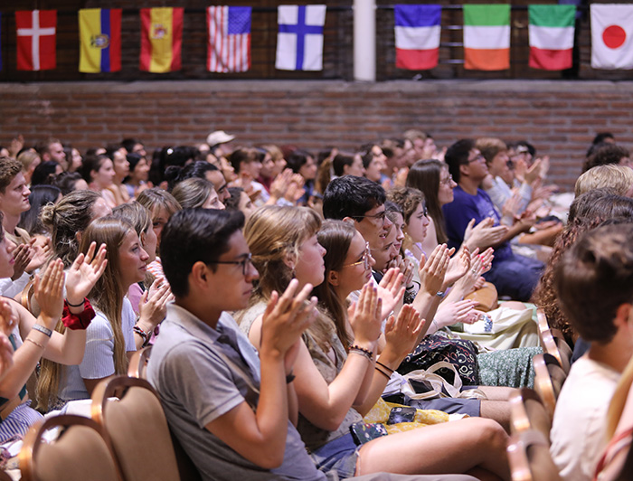 A group of international students are clapping. In the background is a wall with flags from Denmark, Colombia, Spain, United States, Finland, France, Ireland, Italy and Japan.