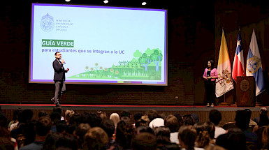 A man is giving a speech on stage while a woman is looking at him waiting to talk. In the wall is a screen with the phrase "Green Guide: for students that are joining to the UC Chile".
