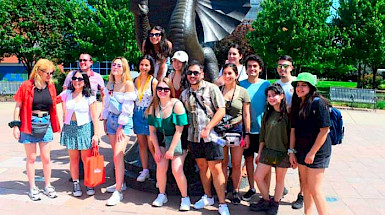 Fourteen students and one instructor are posing for the photo in front of a dragon statue, the Drexel University symbol.