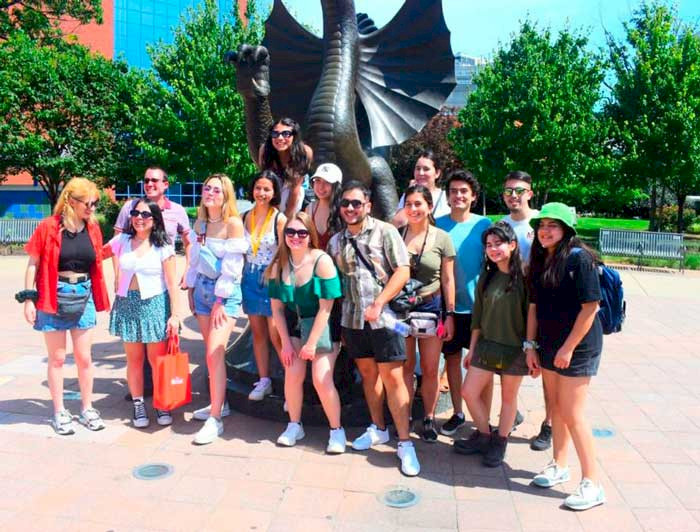 Fourteen students and one instructor are posing for the photo in front of a dragon statue, the Drexel University symbol.