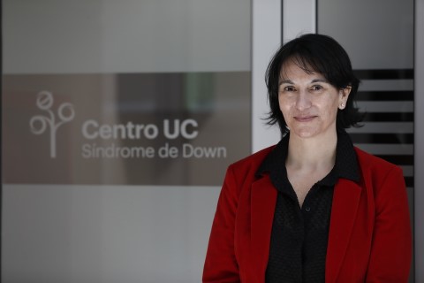 Macarena Lizama is posing with a smile on her face in front of the UC Chile Down Syndrome Center. She is dressed with a black blouse and a red blazer. 